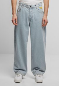 Lyseblå vide jeans med avslappet passform, fem lommer og gult logo-detalj foran. Brukt med hvite sneakers.