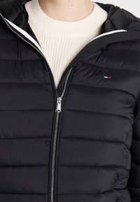 Černá péřová bunda s prošívanými detaily, zapínáním na zip a kapucí. Obsahuje malou logo akcent na hrudi a bílé podšívání.