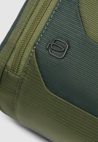 Borsa in tessuto verde con pannello verde scuro, chiusura con zip e dettaglio logo nero.