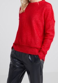 Roter Strickpullover mit lässiger Passform, geripptem Saum und Bündchen, getragen über schwarzen Kunstlederhosen. Weiche Textur mit leichtem Glanz.