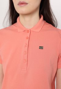 Koralo spalvos polo marškiniai su klasikinėmis apykaklėmis, penkių mygtukų užsegimu ir mažuoju juodu logotipu kairėje pusėje. Pagaminti iš tekstūruoto medvilninio audinio.