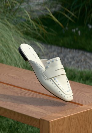 Hvid slip-on sko med sølvnitter flydende over en træ bænk udendørs med græs og grus i baggrunden.