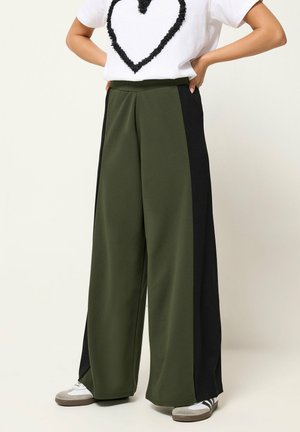 Pantaloni a gamba larga verde oliva con pannelli laterali neri, indossati con sneakers bianche, abbinati a una maglietta bianca con motivo a cuore nero.