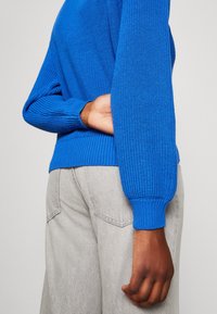 Pull en tricot bleu avec poignets et ourlet côtelés, présentant une coupe ample. Porté avec un pantalon gris clair, mettant en valeur les détails du tissu texturé.