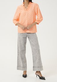 Koralfarvet bluse med zigzagmønster og flæsekant ved halsen, 3/4-ærmer, kombineret med højtaljede, lysegrå vide jeans og sorte mulehøje hæle.