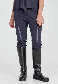 Námornícke cargo nohavice vyrobené z ľahkej tkaniny, s viacerými vreckami na zips a pásom na sťahovanie, v kombinácii s vysokými čiernymi gumákmi.