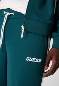 Turquoise trainingsbroek met witte accenten en een trekkoord. Kenmerkt zich door een gestructureerde stof en het 'GUESS' logo in het wit op het been. Ritssluiting zichtbaar.
