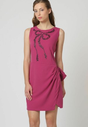Joven mujer con vestido fucsia sin mangas con diseño de lazo de cuentas negras en el pecho y detalle de tela fruncida en la cadera derecha.