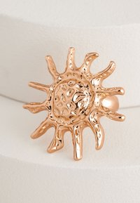 Ring featuring a textured, abstract sun design in rose gold metal. Rounded central disc with pointed rays, polished surface and varied thickness.