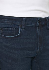 Primer plano de la cintura y los bolsillos de unos pantalones de mezclilla azul oscuro, combinados con una camiseta blanca de canalé metida por dentro.