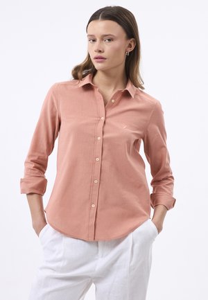 Vrouw draagt een stoffig roze overhemd met knopen en opgerolde mouwen en witte broek, staand met handen in de zakken tegen een effen achtergrond.