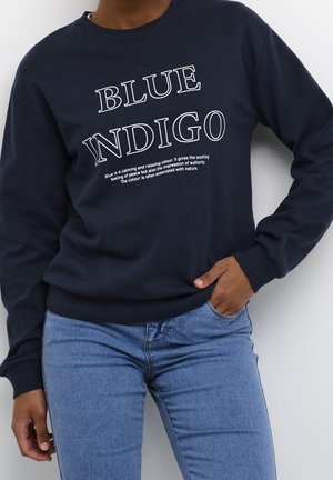 Person iført navyblå sweatshirt med "BLUE INDIGO" og beskrivende tekst, kombineret med lyseblå jeans, stående mod en hvid væg.