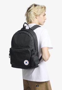 Černý nylonový batoh s kulatým tvarem, vybavený několika přihrádkami, předním zipovým kapsou a nášivkou s logem Converse All Star.