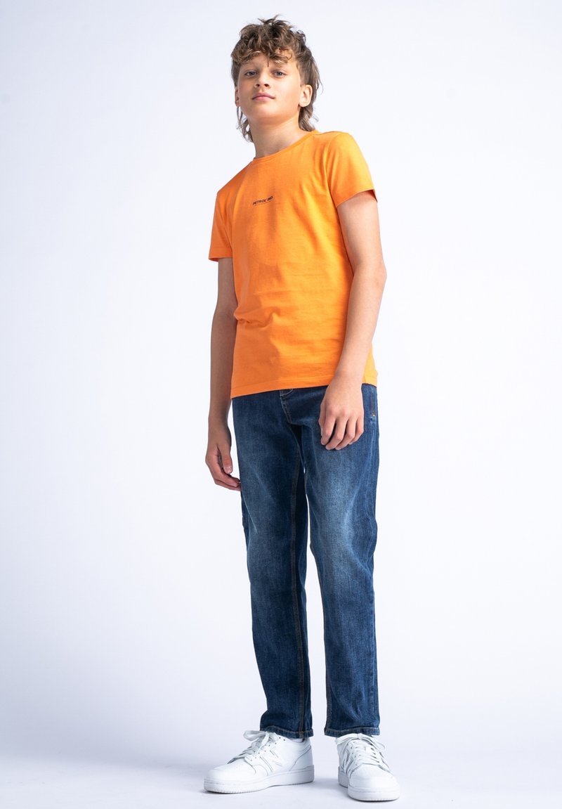 Camiseta de algodón naranja de manga corta, combinada con jeans de mezclilla azules. Zapatillas blancas completan el conjunto. Diseño simple y casual.
