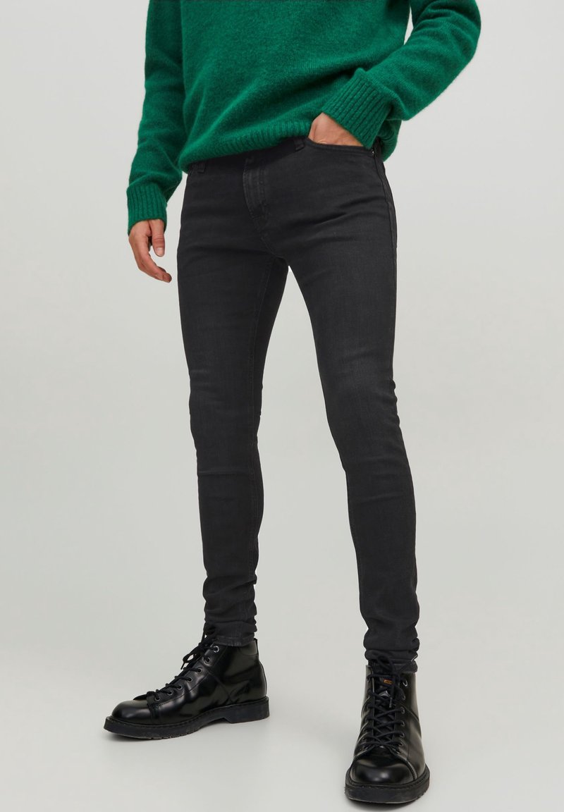 Jeans de mezclilla negros de corte slim con una textura suave, diseño clásico de cinco bolsillos y sin desgastes visibles. Se llevan con botas de combate negras.