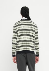 Persoon met gevlochten haar, gekleed in een zwart-wit geblokt sweater en donkere broek, met de rug naar de camera tegen een effen witte achtergrond.