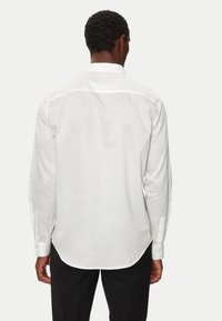 Camicia bianca a maniche lunghe con colletto alla cinese, taglio dritto, texture liscia e polsini abbottonati. La vista posteriore mostra cuciture sulle spalle sottili.