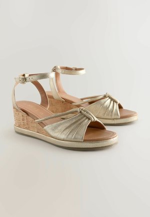 Auksiniai kūgio formos sandalai su kamštiniu kulnu, dekoruotomis dirželių detalėmis, suknoru priekyje ir kulkšnies užsegimu. Minkšta vidinė pamušalas ir tekstūruotas padas geresniam sukibimui.