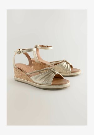 Goldene Keilsandalen mit Korkabsatz, verzierten Riemen, geknoteter Vorderseite und Knöchelriemen. Weiche Innenauskleidung und strukturierte Sohle für besseren Halt.