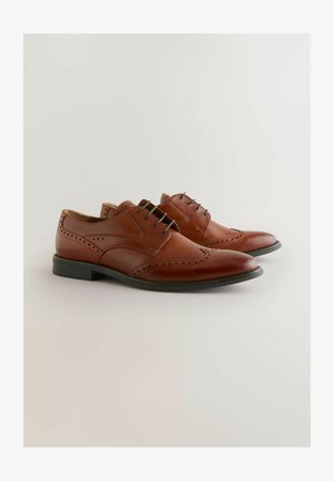 Ein Paar braune Lederbrogue-Schnürschuhe mit durchbrochener Detailgestaltung und schwarzen Sohlen vor einem schlichten hellen Hintergrund.