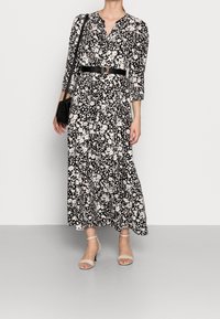Robe longue noire et blanche à motifs floraux avec des manches trois-quarts, un col en V, une taille cintrée avec une ceinture noire et une silhouette fluide.