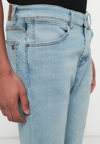 Světle modré džínové kalhoty s klasickým designem pěti kapes, zdobené proužkováním a hnědou koženou nášivkou s logem na zadním pasu.