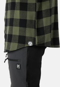 Grün und schwarz karierte Flanellhemd mit einem Etikett, kombiniert mit schwarzen Hosen mit einer Reißverschlusstasche und einem X-Trail-Logo.