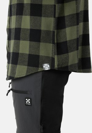 Grünes und schwarzes kariertes Flanellhemd mit einem Etikett, kombiniert mit schwarzen Hosen, die eine Reißverschlusstasche und einen Logoschriftzug auf dem Oberschenkel haben.