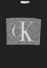 Černý svetr s kulatým výstřihem a středovým šedým obdélníkovým pruhem. Uprostřed je výrazně umístěno bílé logo "CK".