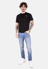 Camiseta negra de manga corta con logo en el pecho, combinada con vaqueros de mezclilla de color azul claro y zapatillas blancas. El modelo tiene tatuajes y el cabello rizado.