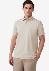 Polo shirt beige a maniche corte con colletto, caratterizzato da un logo discreto sul petto e un tessuto morbido e leggero. Abbinato a pantaloni di colore chiaro.
