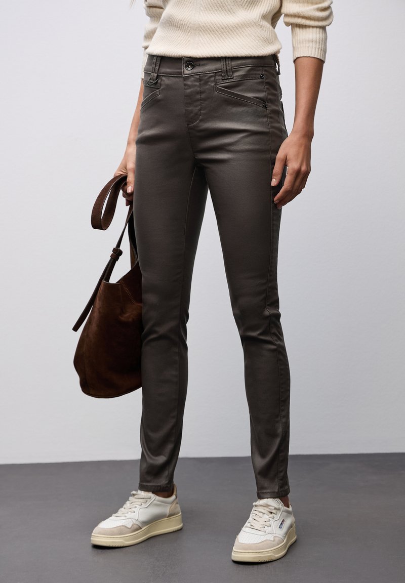 Pantalones ajustados de color marrón oscuro hechos de una tela suave, con bolsillos frontales y cremallera, combinados con un bolso de ante marrón y zapatillas blancas.