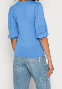 Světle modrý top s nabíranými rukávy, kulatým výstřihem a nařasenými manžetami. V kombinaci se světle modrými džíny s detaily v podobě kapek barvy.