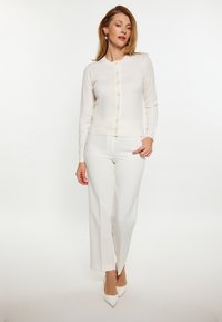 Leichter, cremefarbener Cardigan mit Rundhalsausschnitt und Knopfverschluss, kombiniert mit weißen, weiten Hosen und spitzen Absatzschuhen.