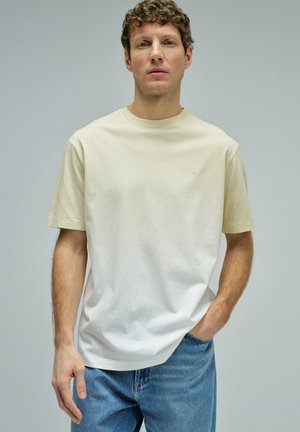 Joven con cabello rizado que lleva una camiseta con un degradado de beige a blanco y vaqueros azules, de pie frente a un fondo gris liso.