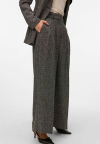 Kobieta w szarych, szerokich spodniach dzwonowych z tkaniny tweed, z kieszeniami, zestawionych z dopasowaną marynarką i czarnymi szpilkami z ostrym noskiem.