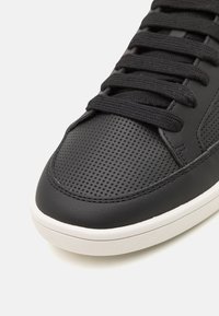 Schwarze Sneaker mit perforierter Textur, flachen Schnürsenkeln und weißer Sohle. Glatte Akzente aus schwarzem Leder und kontrastierende Nähte.