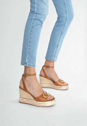 Jambes portant un jean skinny bleu clair et des sandales compensées marron avec des brides à la cheville et des boucles dorées sur fond blanc.