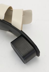 Sandale à bride synthétique crème avec un talon bloc noir arrondi, présentant une texture lisse et un design minimaliste. Notable pour ses lignes épurées.