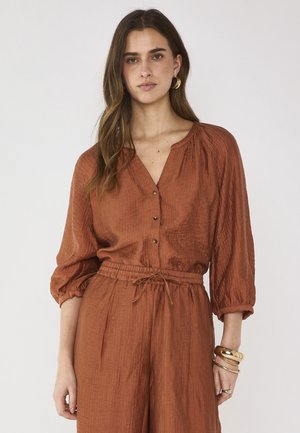 Vrouw met lang bruin haar, gekleed in een roestkleurige bloes met knopen en driekwart mouwen, bijpassende broek met aantrekkoord, gouden creolen en armbanden.