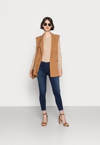Blazer marrón sin mangas, top de canalé beige, jeans ajustados azul oscuro y sandalias de tacón marrones. El modelo también lleva gafas de sol redondas negras.