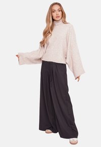 Jasnoróżowy sweter o grubym splotach z wysokim kołnierzem, zestawiony z czarnymi spodniami o szerokich nogawkach i beżowymi trampkami na platformie. Miękka struktura, luźny fason.