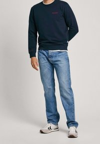 Sudadera navy con logo rojo, combinada con jeans de pierna recta azul claro y zapatillas blancas con detalles en negro. Diseño simple y casual.