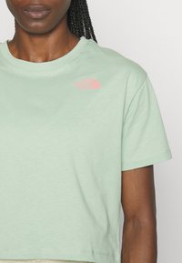 Světle zelené bavlněné tričko s krátkými rukávy a kulatým výstřihem, s růžovým logem na levé straně hrudi. Hladká textura.