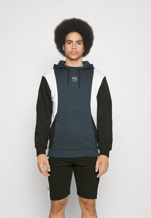Kapuzensweatshirt mit einem türkisfarbenen Körper, schwarzen Ärmeln und weißen Seitenelementen. Mit einem Logo, Kordelzug und gerippten Bündchen. Kombiniert mit schwarzen Shorts.