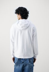 Hvit hettegenser med avslappet passform, laget av myk stoff, med kengurulomme og ribbestrikkede mansjetter, kombinert med blå jeans.
