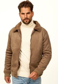 Braune Wildlederjacke mit einem flauschigen, strukturierten Kragen. Verfügt über einen Reißverschluss und gerippte Bündchen, getragen über einem cremefarbenen Rollkragenpullover und hellen Jeans.