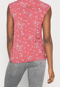 Haut sans manches fleuri en tissu rose avec une coupe ample. Présente de petits motifs de papillons bleus et blancs et un dos plissé.