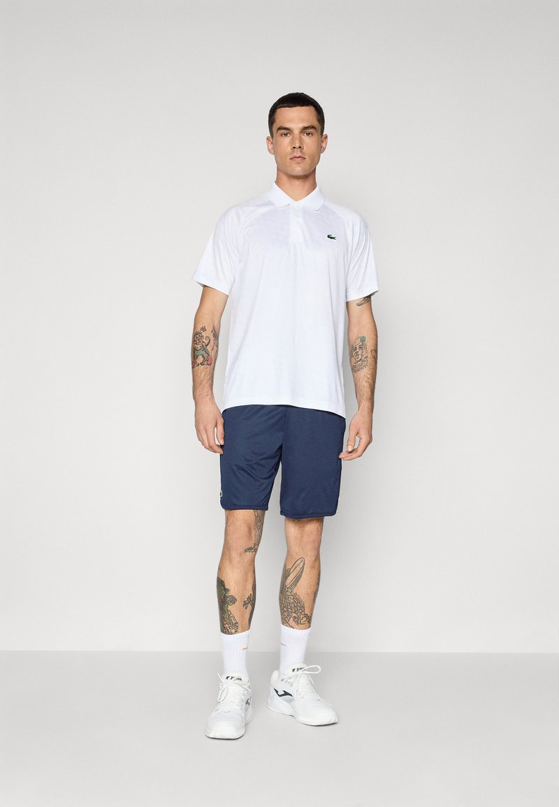 Polo shirt blanc à manches courtes avec un logo vert, associé à un short bleu marine. Modèle portant des baskets blanches et des chaussettes mi-mollet, affichant des tatouages.