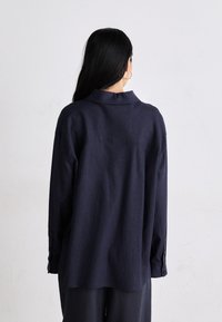 Camisa de manga larga azul marino fabricada en tejido ligero, con un cuello suave, dobladillo curvado y puños abotonados. Diseño simple y de ajuste relajado.
